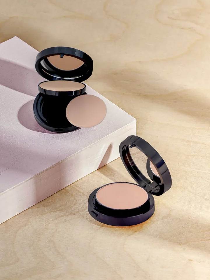 Two open and one closed compact foundation/powder cases on a light pink block and wooden surface, showing neutral beige tones.