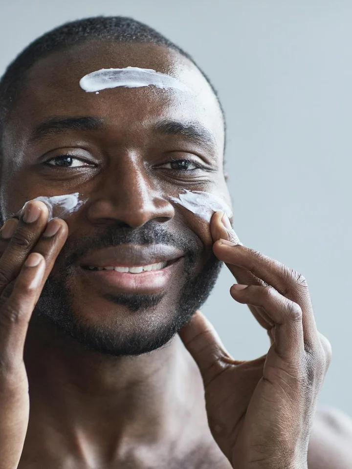Close-up of a smiling man applying a white cream to his face for a skincare routine, illustrating moisturizer use.
