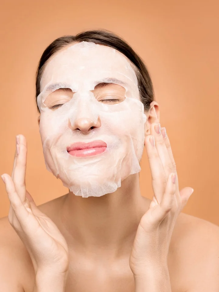 Woman wearing a white sheet mask with eyes closed, hands raised near her face.