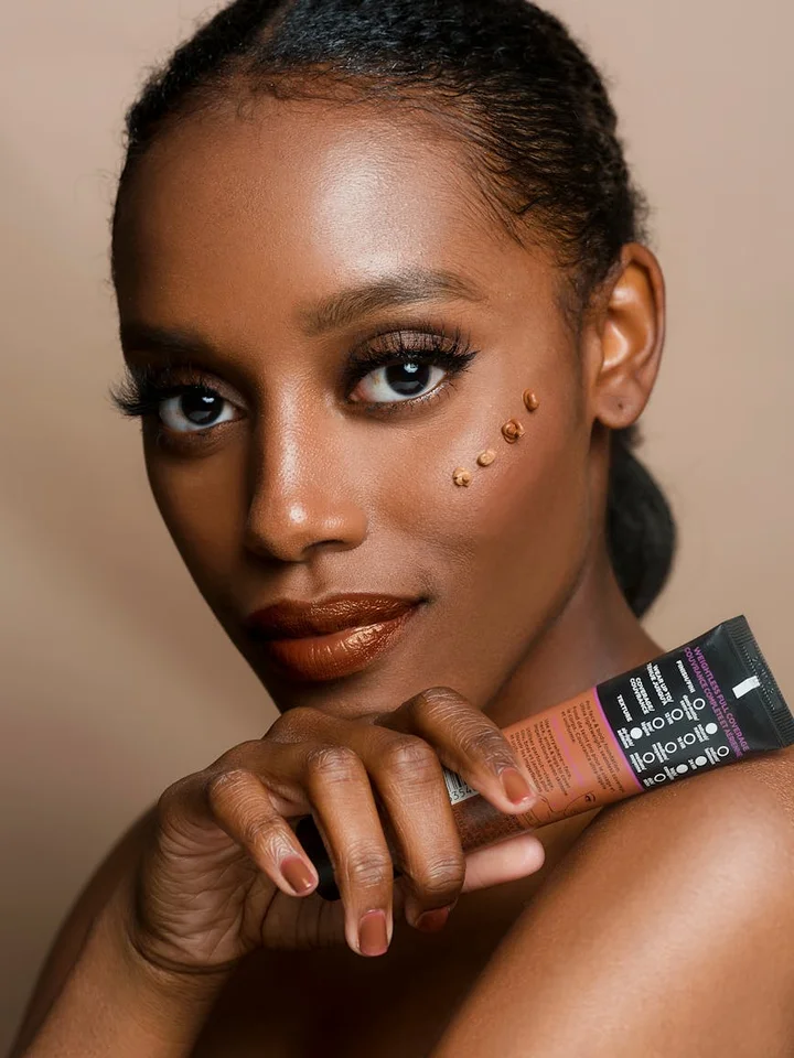 Close-up portrait of a woman holding a foundation tube near her face, with decorative dots on her cheek and glossy makeup.