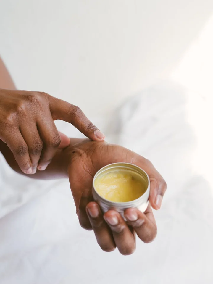 Close-up of hands applying a small jar of moisturizer onto the back of one hand with a soft white background.