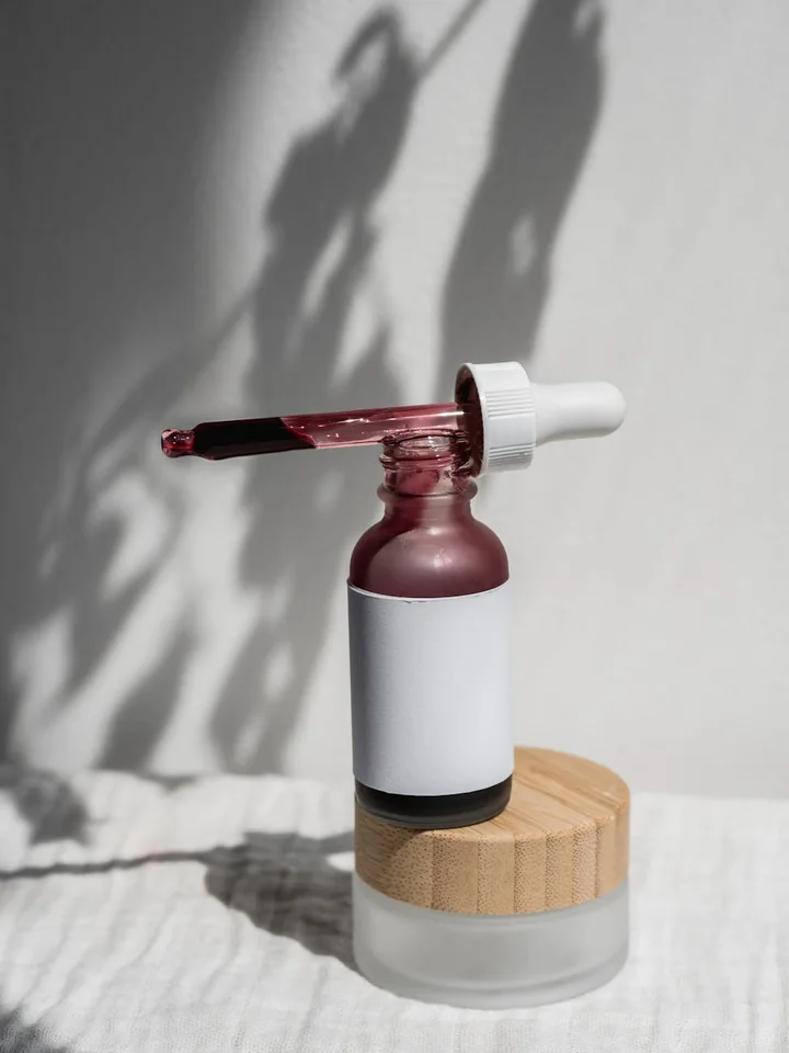 Close-up of a dropper bottle with pink serum and a white cap resting above a wooden-cap jar on a white surface.