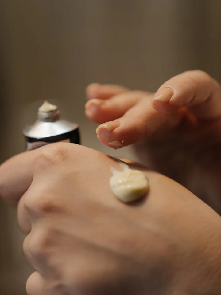 Close-up of a dollop of cream on the back of a hand with a skincare tube in the background.
