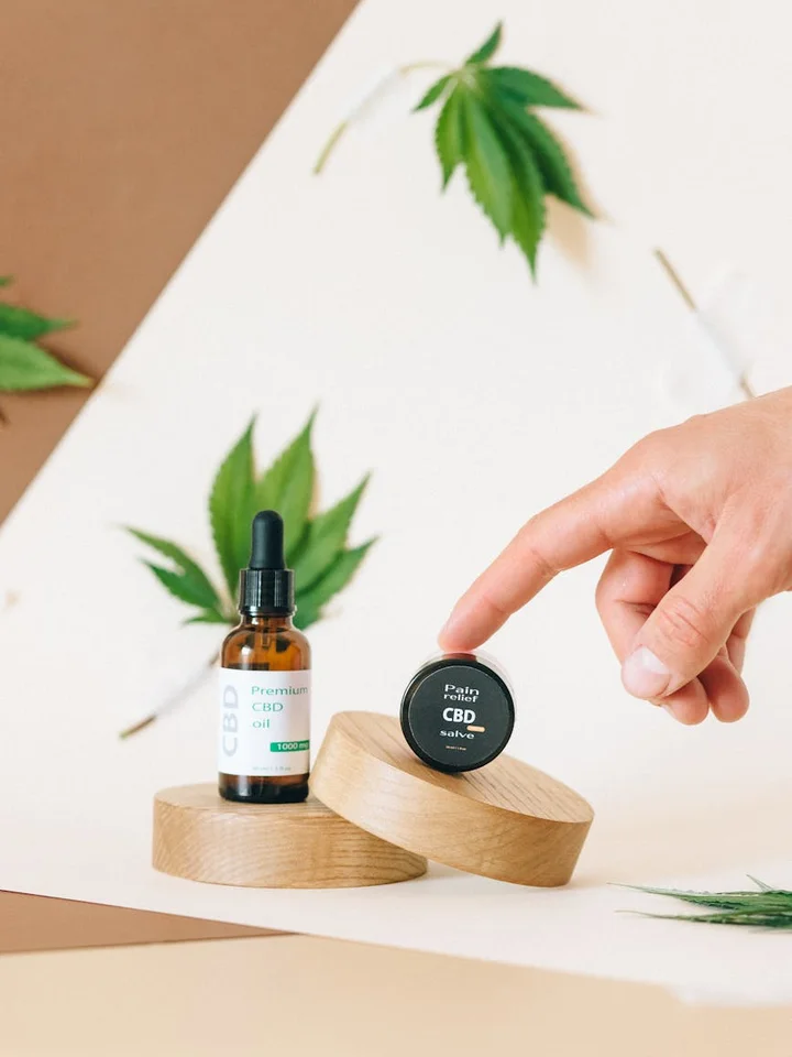 A bottle of CBD oil and a small jar on wooden pedestals, with green leaves in the background.