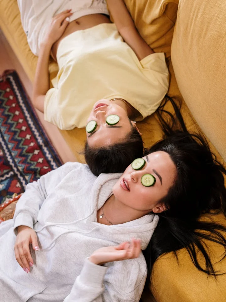 Two women relaxing on a couch with cucumber slices over their eyes during a spa treatment, wearing robes.