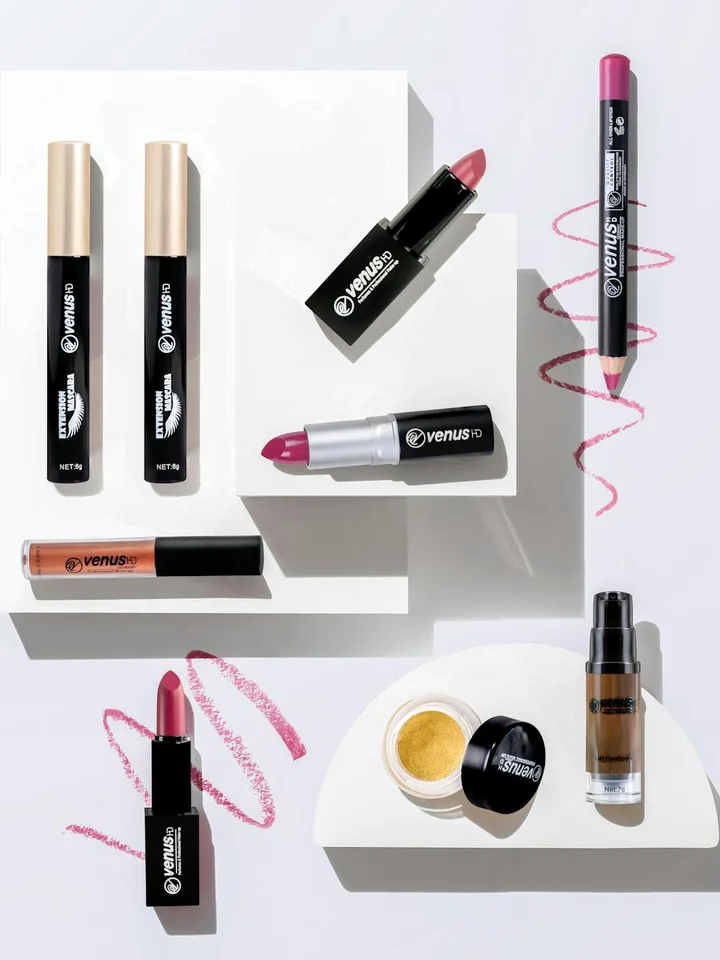 Flat lay of NYX cosmetics including lipsticks, lip liners, a compact powder, and a liquid foundation bottle on white geometric platforms with pink scribble lines in the background.