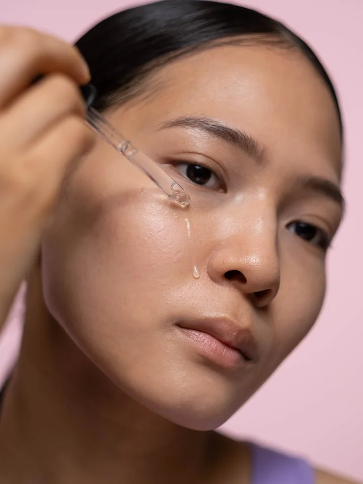 A person applies hyaluronic acid serum to their face using a dropper, with a pink background.