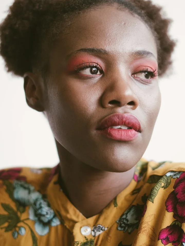 Close-up portrait of a Black woman with pink eyeshadow and pink lipstick, wearing a yellow floral blouse.
