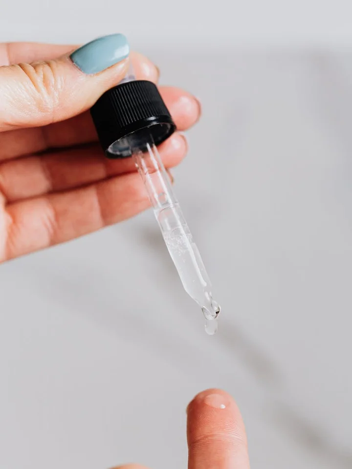 Close-up of a skincare dropper with clear liquid about to be applied to the skin; hand with blue-nail polish holds the dropper.