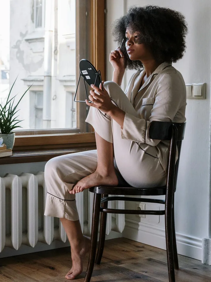 Woman sitting by a window applying skincare with a handheld mirror, bathed in natural morning light.