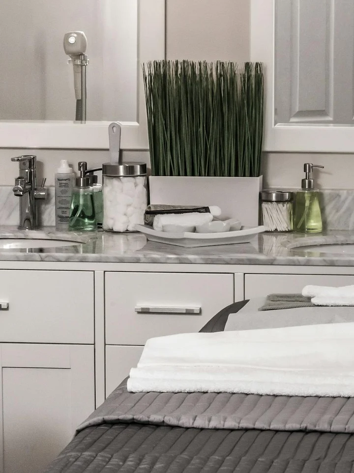 Spa-style bathroom countertop with skincare products, a tall green plant in a white pot, and folded towels, against white cabinets and a marble surface.