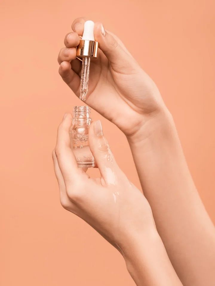 Close-up of hands holding a glass dropper bottle and applying serum on the skin against a peach-colored background.