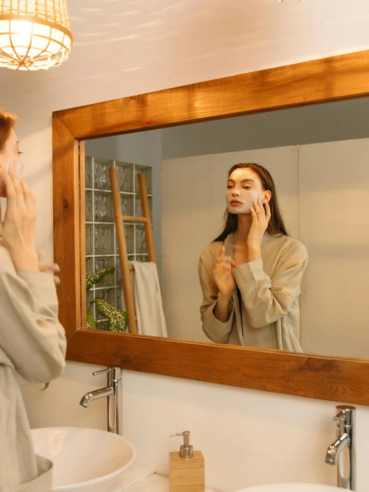 Person in a robe applying skincare product in front of a bathroom mirror