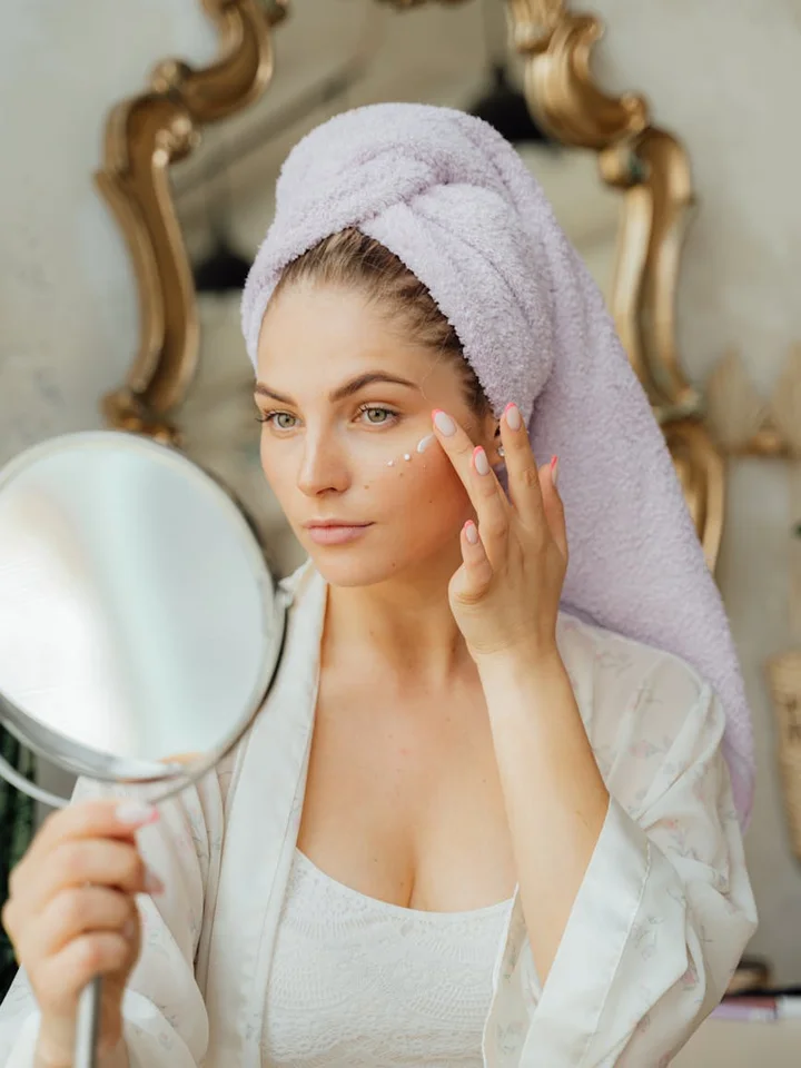 Woman with a towel wrapped around her head applying skincare in front of a mirror.