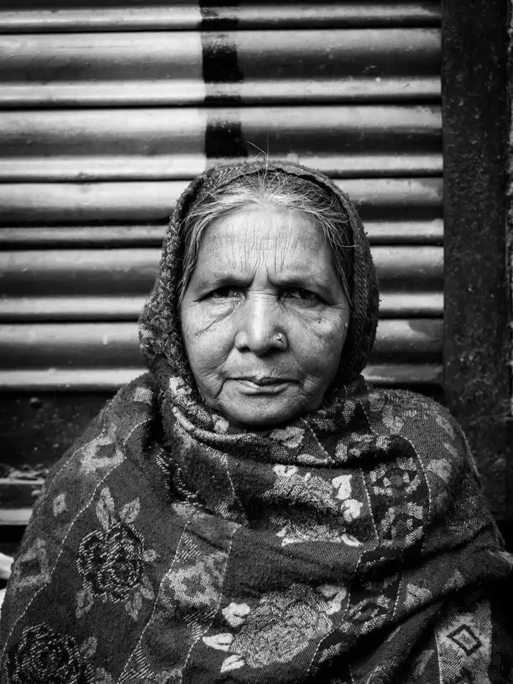 Black-and-white portrait of an older woman wearing a patterned shawl, standing in front of a striped background.
