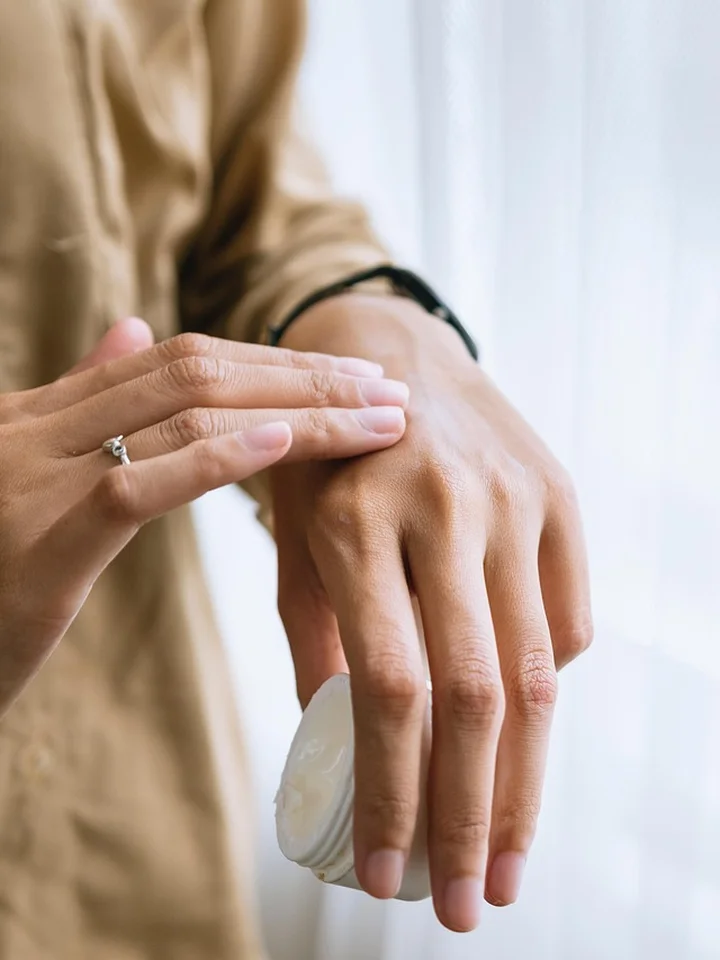 Close-up of a person applying skincare product from a small jar onto the back of their hand, with a ring visible on the other hand.