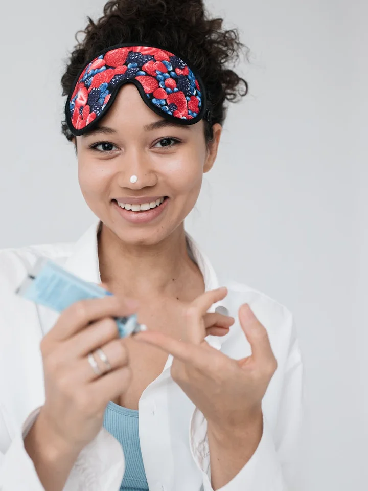 Smiling woman wearing a red-patterned sleep mask applies a skincare product under her eyes.