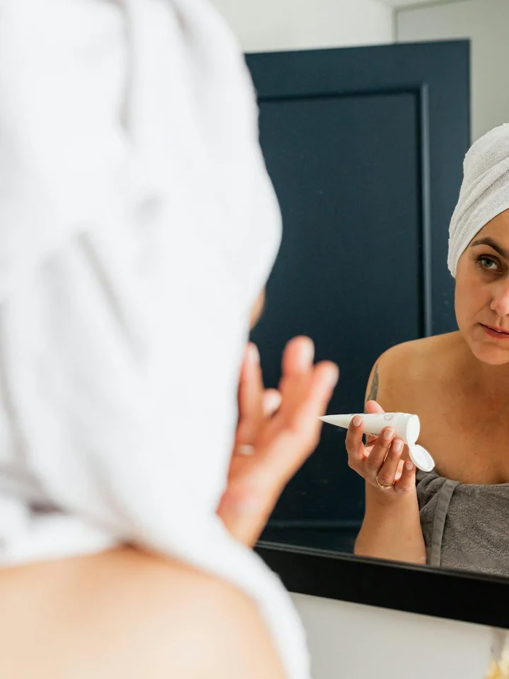 Person with a towel on their head looking into a bathroom mirror while holding a skincare tube.
