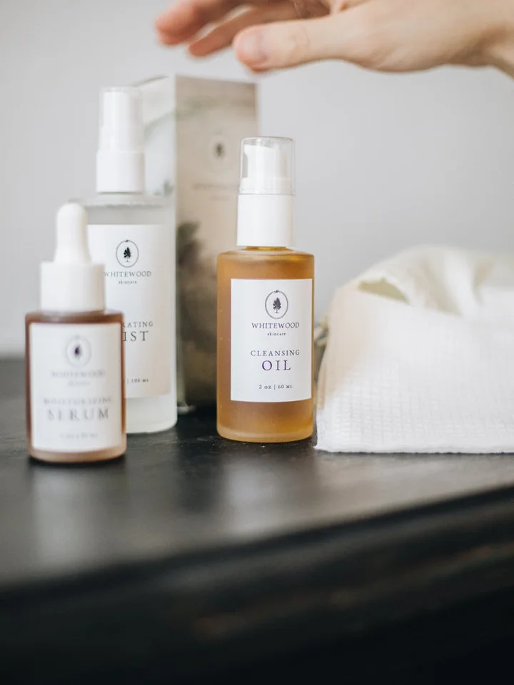 A hand reaching toward skincare bottles on a dark surface, including a cleansing oil and serum, with a white towel in the background.