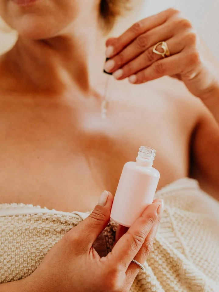 Close-up of a person applying skincare serum from a white bottle, with a towel draped over the shoulders.