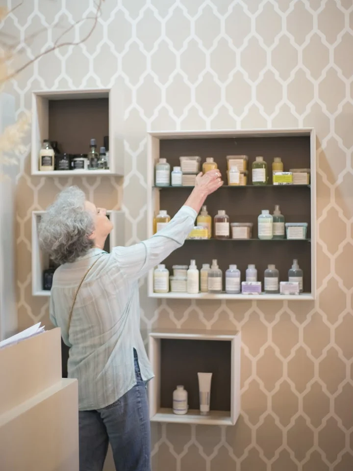 Older person with gray hair reaching for skincare bottles on wall-mounted shelves against a beige geometric-patterned wall.