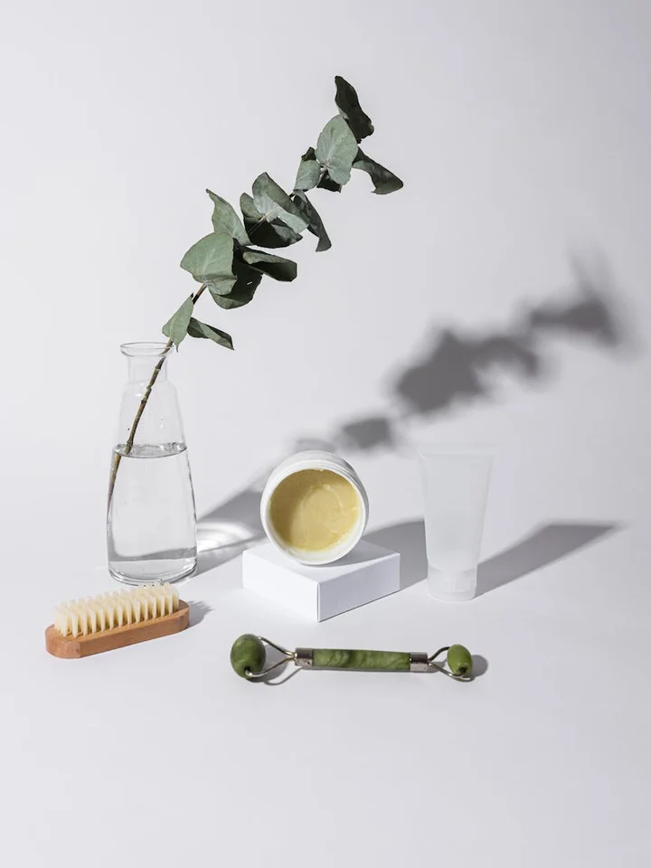 Minimalist skincare scene with a sprig of eucalyptus in a small glass bottle, a jar of cream on a white block, a wooden brush, a glass tube, and a jade roller arranged on a white surface.