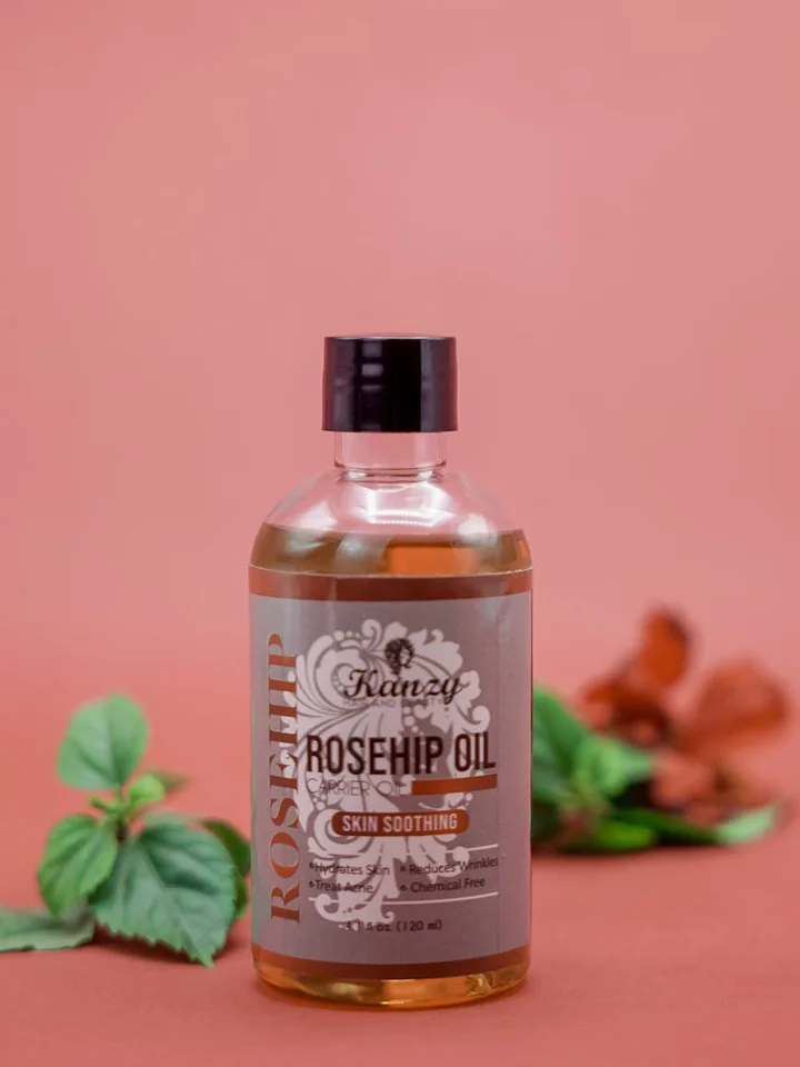 Small bottle labeled Rosehip Oil on a pink background with green leaves nearby.