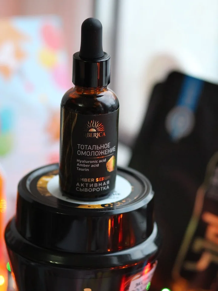 Dark amber dropper bottle with Cyrillic label perched on a black cosmetic jar, with a blurred colorful background