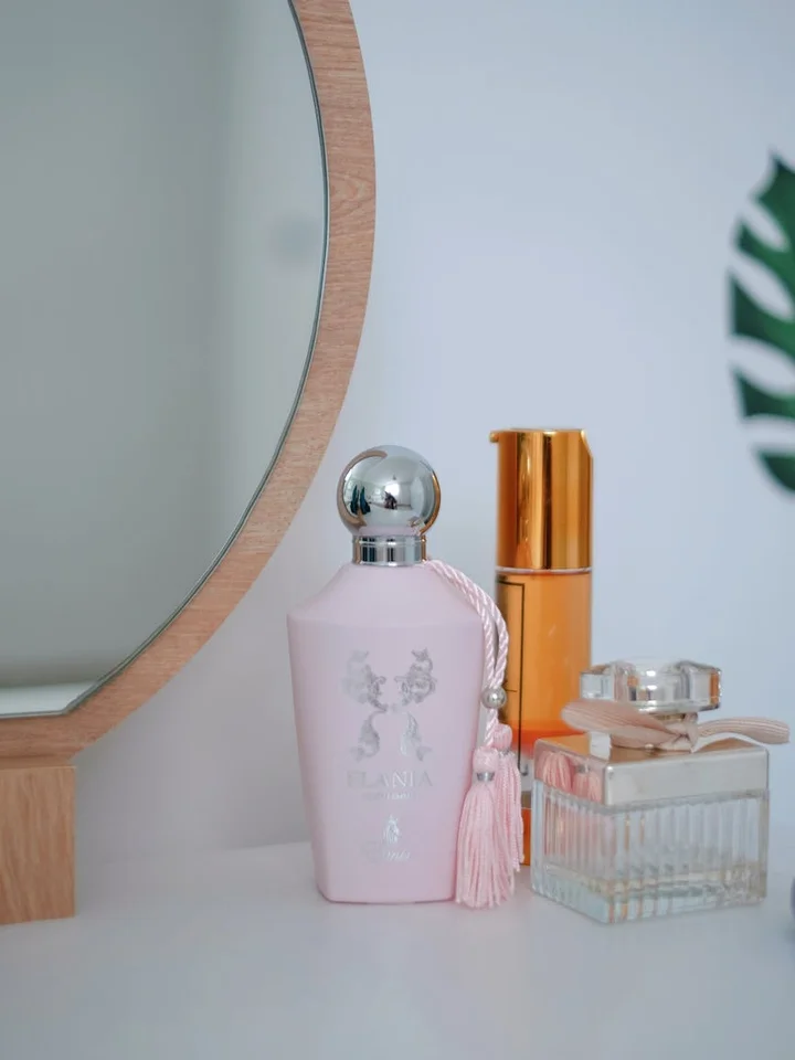 A pink self-tanner bottle with a silver cap and pink tassel, plus clear and amber bottles, arranged on a white surface beside a round wooden-framed mirror.