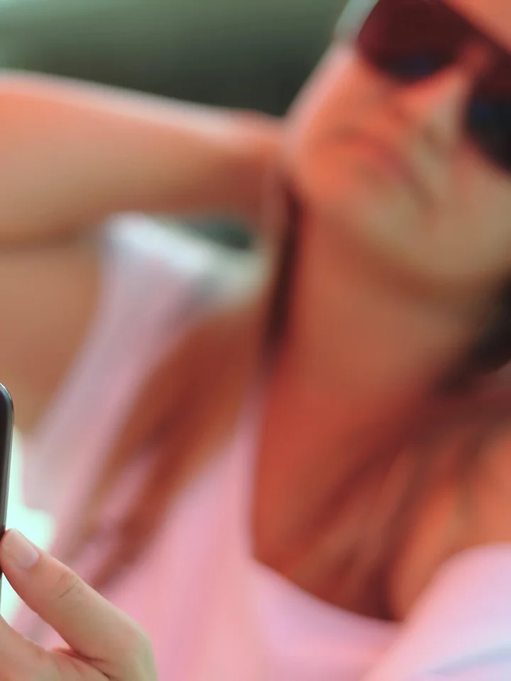 Blurred photo of a person wearing sunglasses and a light pink shirt, holding a smartphone outdoors.