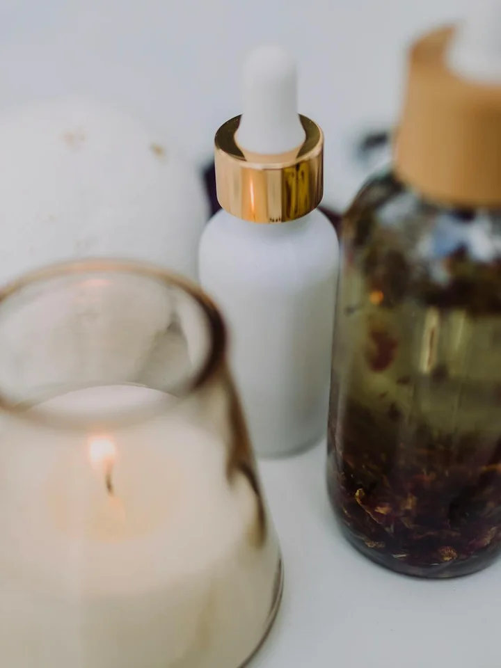 Close-up of skincare bottles on a white surface, including a white dropper bottle with a gold cap and a brown glass bottle, with a blurred candle in the foreground