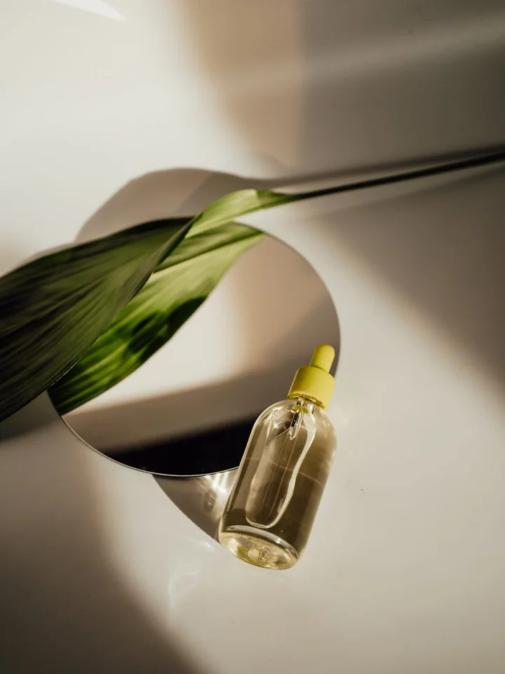 A glass serum bottle with a dropper beside a green leaf, reflected in a round mirror on a clean white surface.