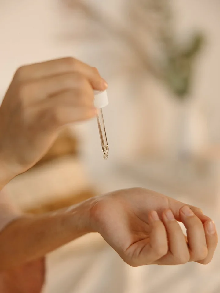 Close-up of a dropper releasing serum onto the back of a hand, with a soft, blurred background.
