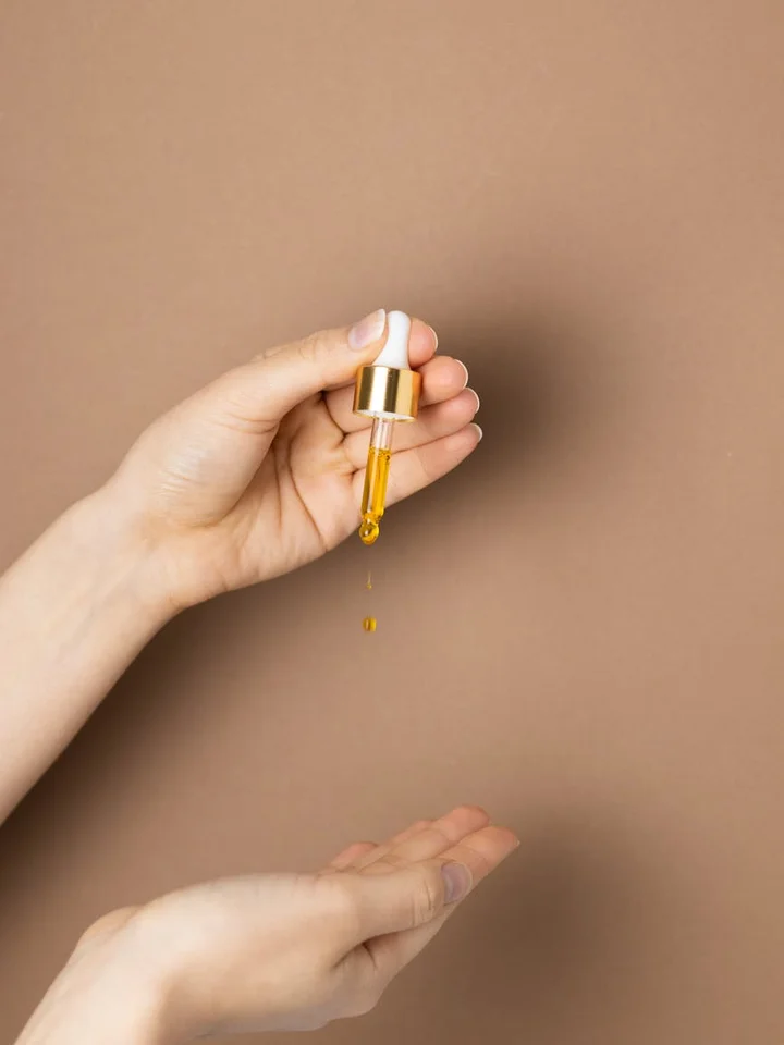 Close-up of a hand holding a dropper with golden serum releasing a few droplets against a beige background.