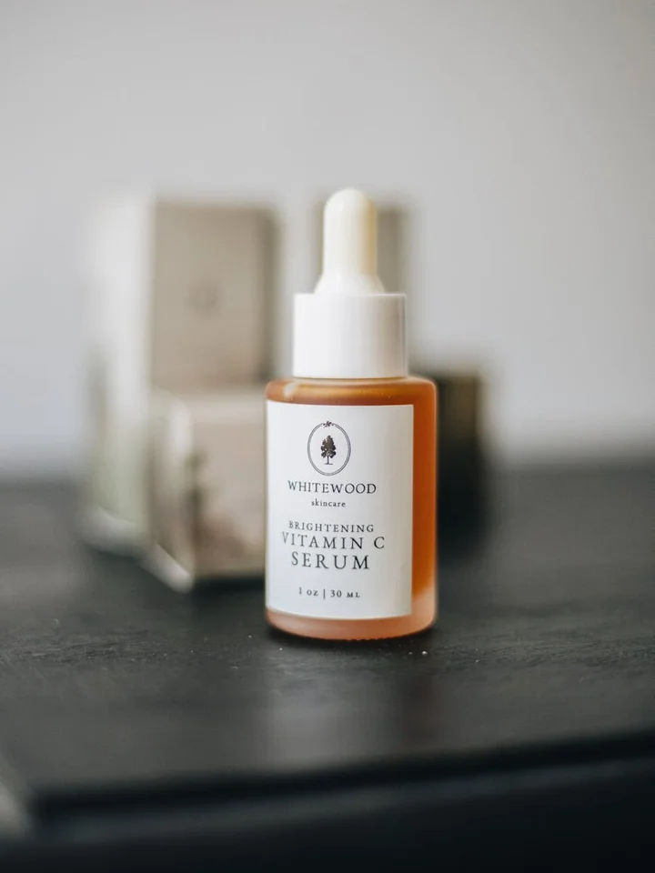 Amber glass dropper bottle labeled Brightening Vitamin C Serum on a dark surface with blurred skincare packaging in the background.