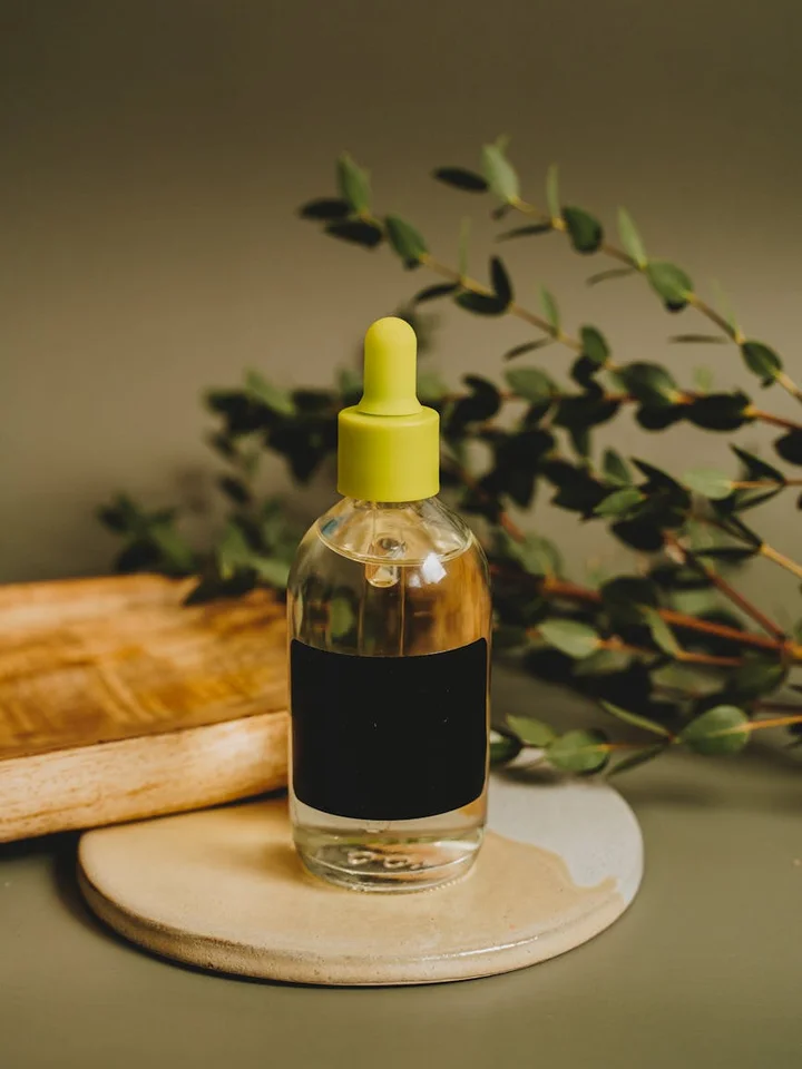 Clear glass serum bottle with a yellow dropper cap on a wooden coaster, with green foliage in the background