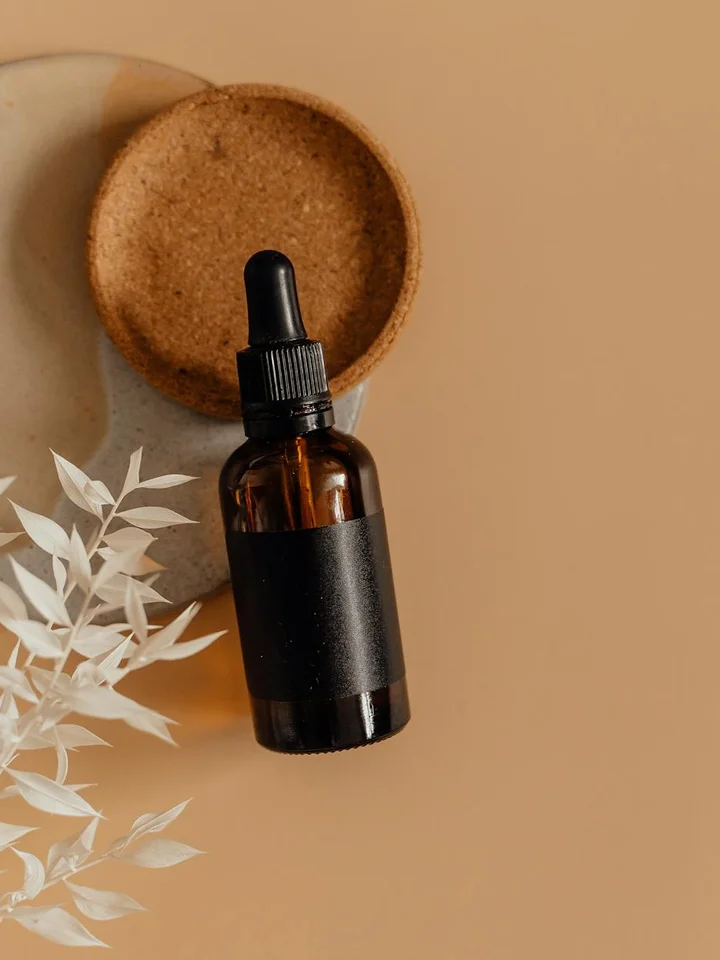 A dark amber glass dropper bottle with a black label sits on a warm beige surface, accompanied by dried white leaves.