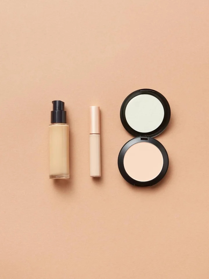 Close-up of a pump foundation bottle, a slender concealer tube, and a round pressed powder compact arranged on a peach background.
