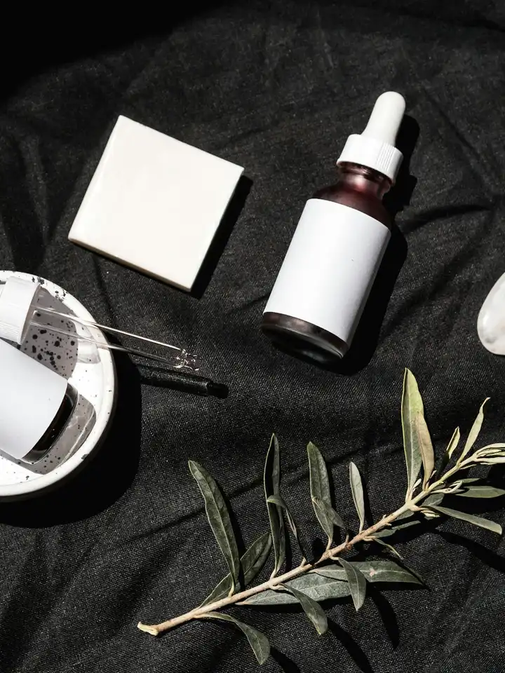 Skincare products on a dark surface, including a dropper bottle, a small white box, and a sprig of greenery.