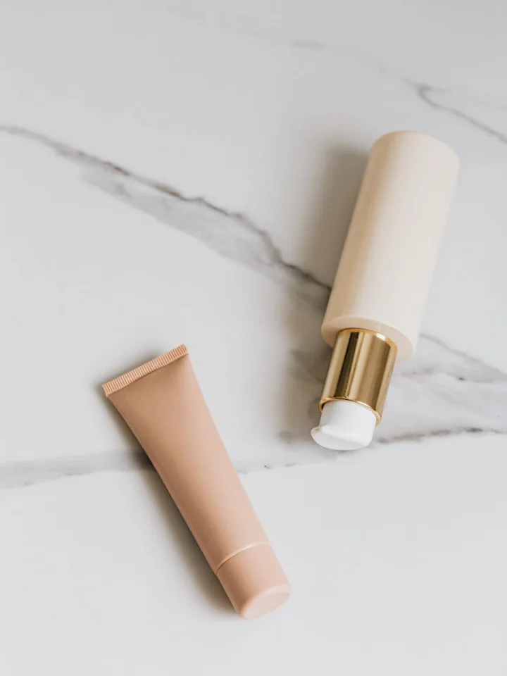 Close-up of two skincare tubes on a white marble surface: a peach-colored squeezable tube and an open-ended bottle with a white applicator exposed.