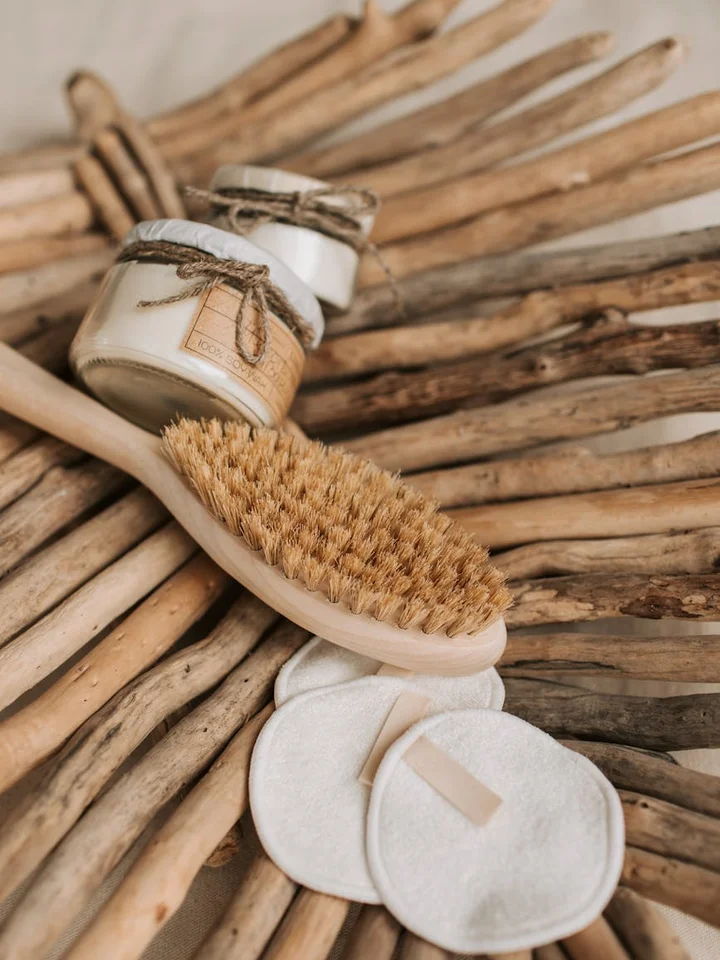 A skincare setup on a woven wooden background featuring a small jar of cream with twine, a natural brush, and white cotton pads.