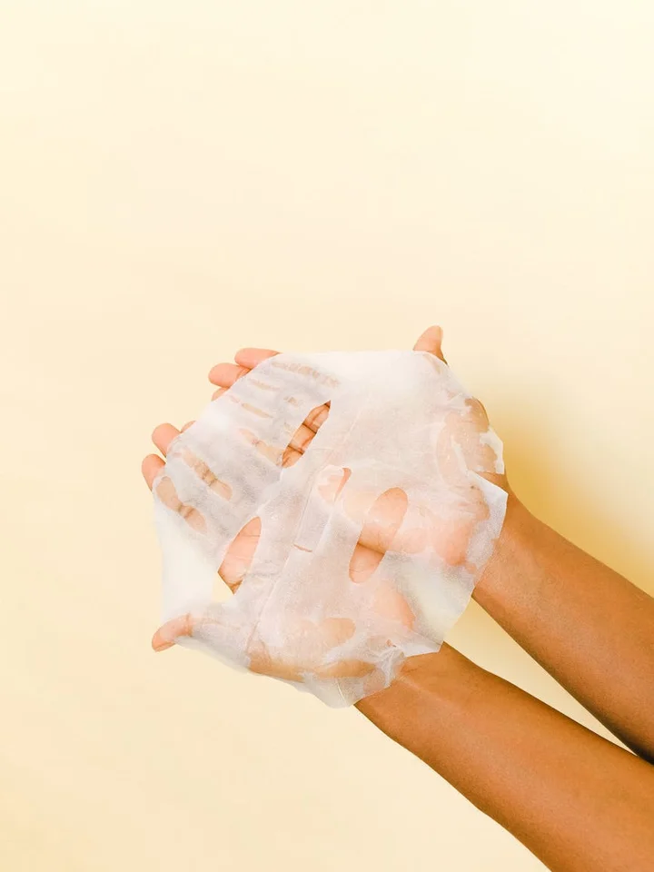 Hands holding a white sheet mask against a pale yellow background