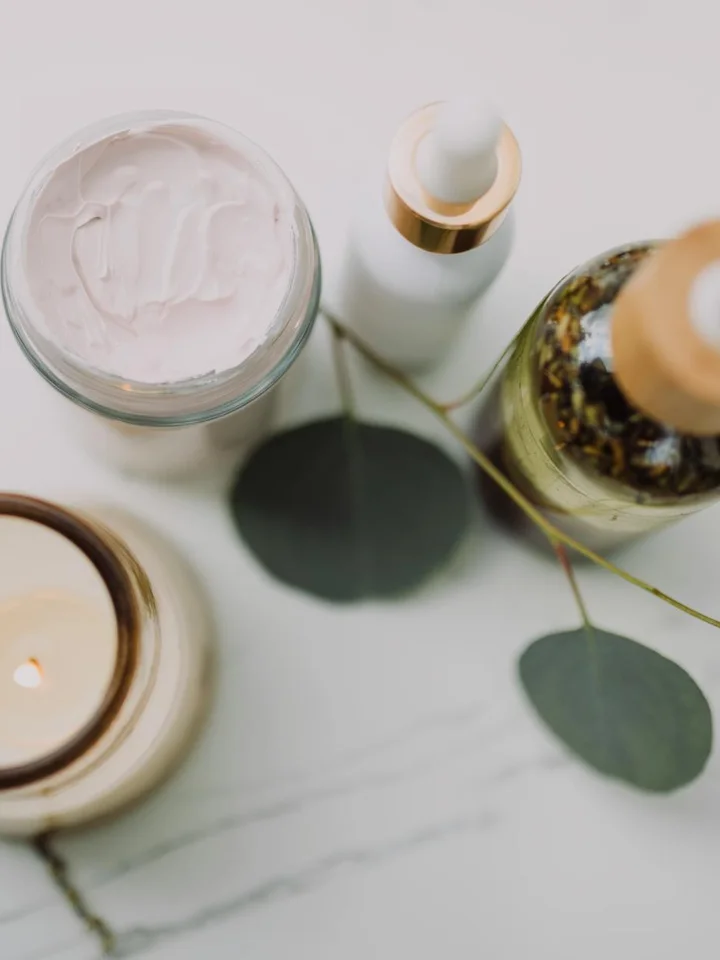 Assorted skincare products arranged on a light marble surface, including a jar of cream, a pump bottle, a dropper bottle, a candle, and eucalyptus leaves.