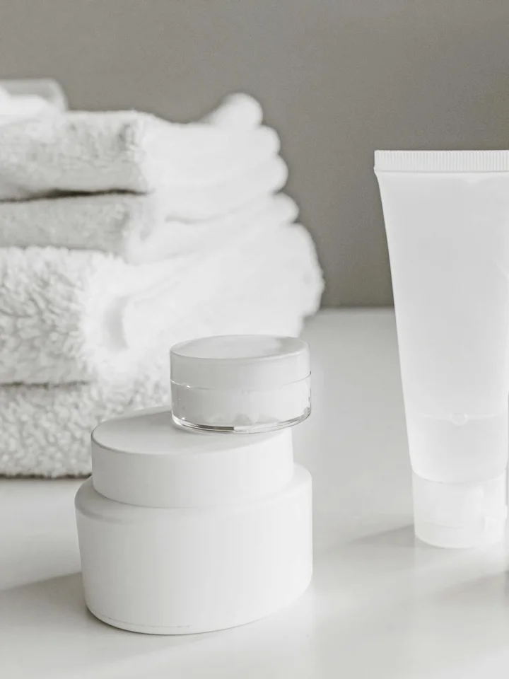 Clean white skincare jars and a tube on a glossy surface with stacked towels in the background