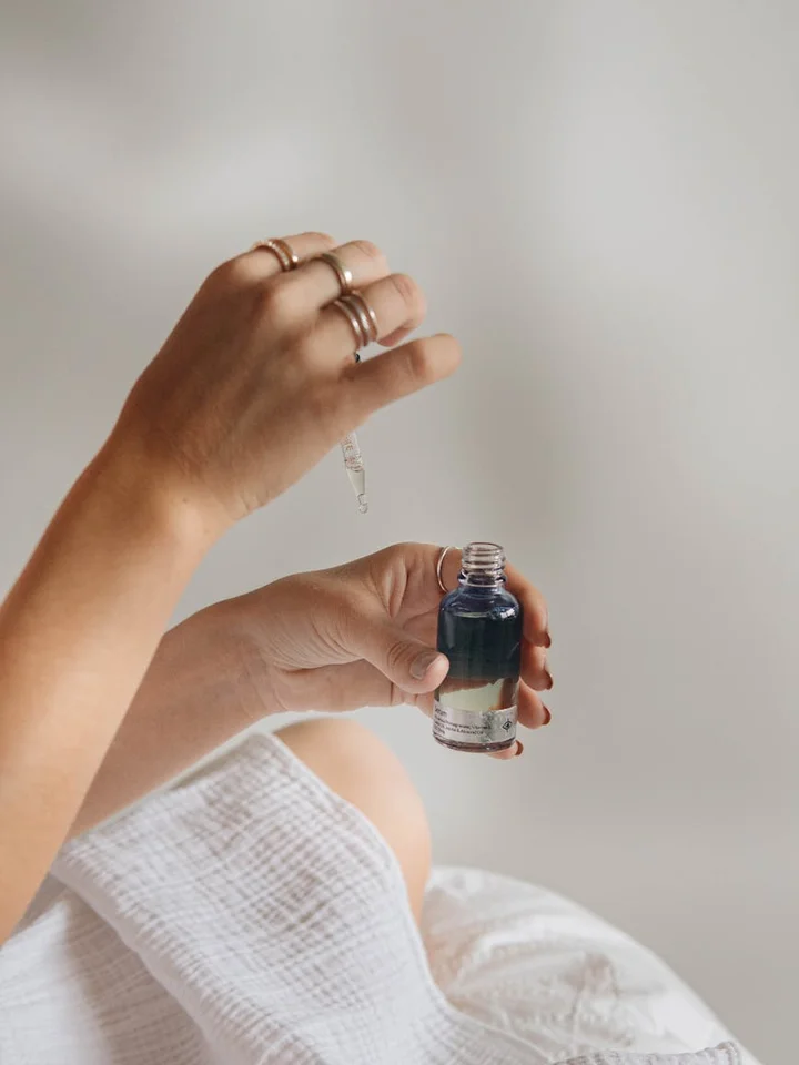 Close-up of a person's hands holding a small dropper bottle with dark blue liquid near a white towel.