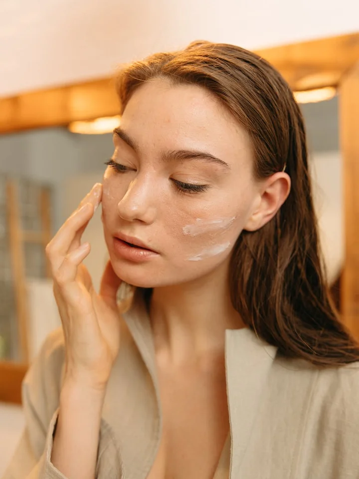 Close-up of a woman applying cream to her face in front of a mirror, as part of a skincare routine.