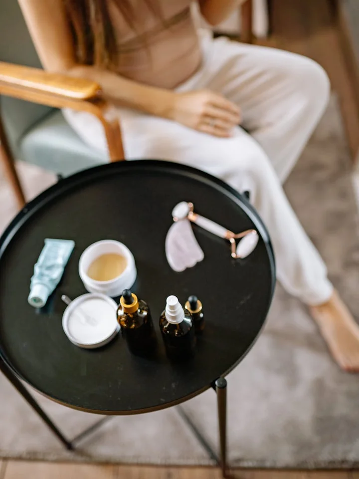 Top-down view of skincare products on a small round table: dropper bottles, a jar, a tube, and a cotton pad, with a person seated in the background.