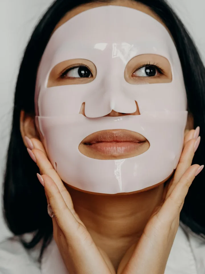 Woman wearing a white sheet facial mask during a skincare routine, with hands framing her face.