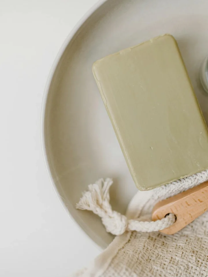 Beige soap bar on a round dish with a loofah and a woven cloth, photographed on a light background.