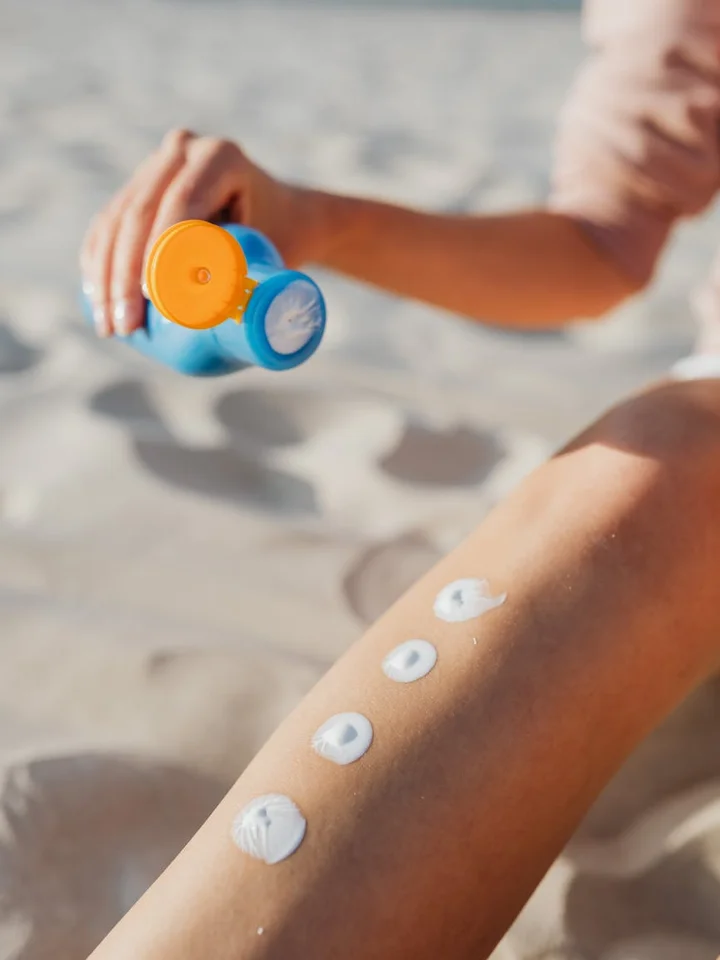 Close-up of a forearm on a sunny beach with several circular sunscreen patches applied, and a sunscreen bottle being squeezed in the background.